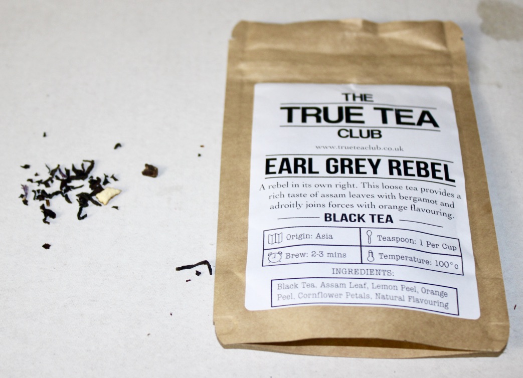 Tea Tastes Terrific | Whittard of Chelsea Apple Crumble Loose Leafed Tea A rectangular brown pouch with a white label that has True Tea Club Earl Grey Rebel Loose Leafed Tea written in black writing on it, with some black Tea Leaves next to the pouch, on a white background.