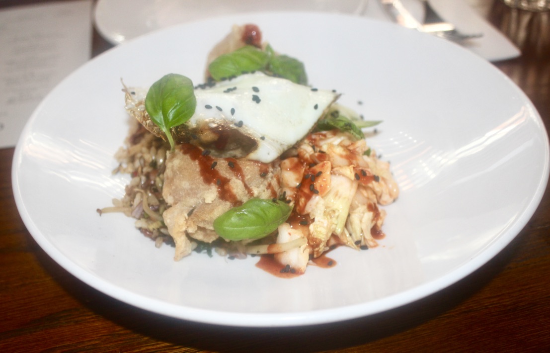 A White Circular plate containing some dark crispy pieces of chicken, some light pieces of Kimchi, and a large white egg with cracked black pepper, on a dark background.