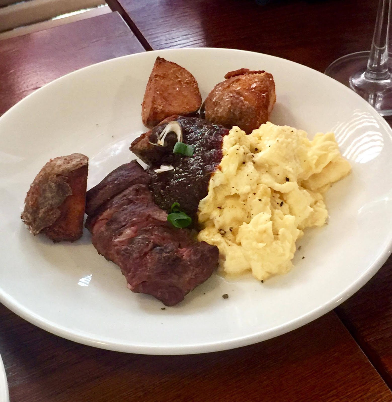A White Circular plate containing some dark pieces of Steak, some light yellow pieces of fluffy Cheddar Eggs, and a large dollops of dark red Chipotle Jam with cracked black pepper, on a dark background.