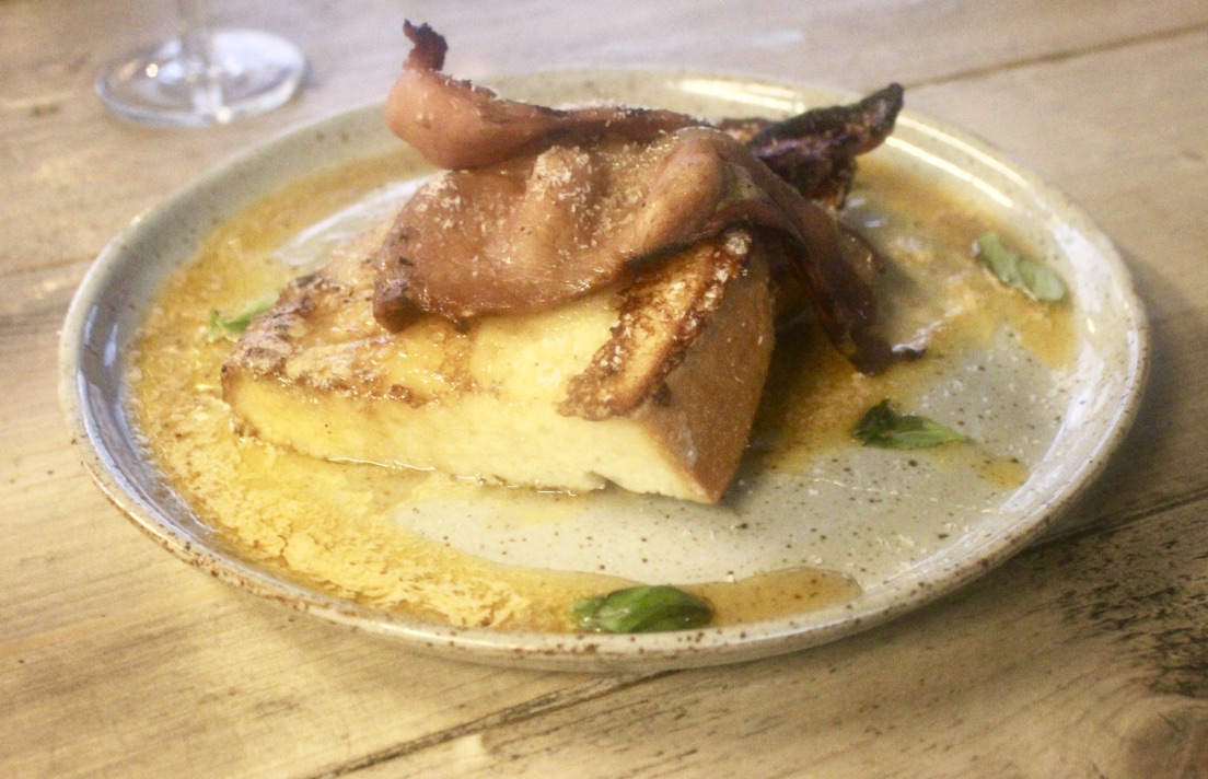 A White Circular plate containing some brightly coloured pieces of Coconut French Toast, covered in a dark coloured butter and topped with some dark brown pieces of crispy Bacon, on a dark background.