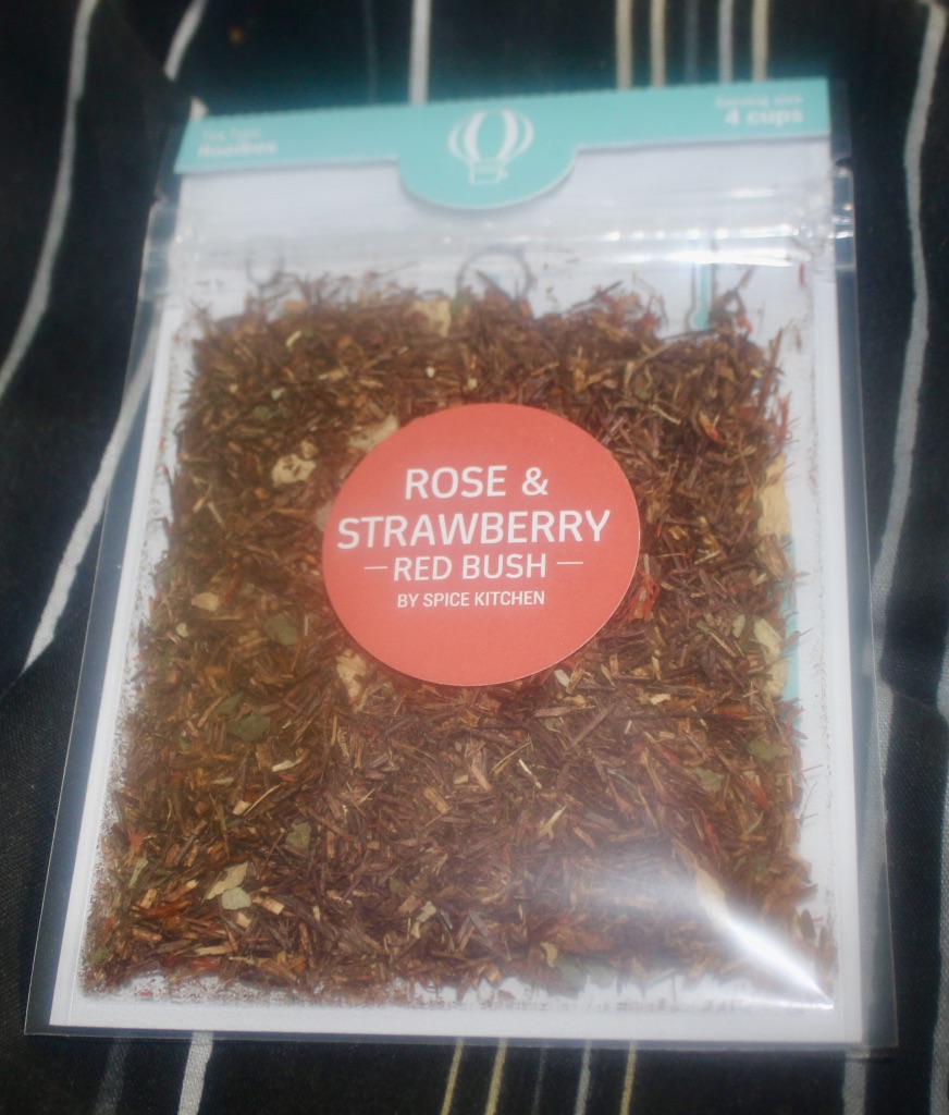 Tea Tastes Terrific | Teatourist March 2017 A silver pouch with a large red circular sticker that has Spice Kitchen UK Rose and Strawberry Rooibos Tea written on it, that contains some dark red tea leaves, on a dark background.