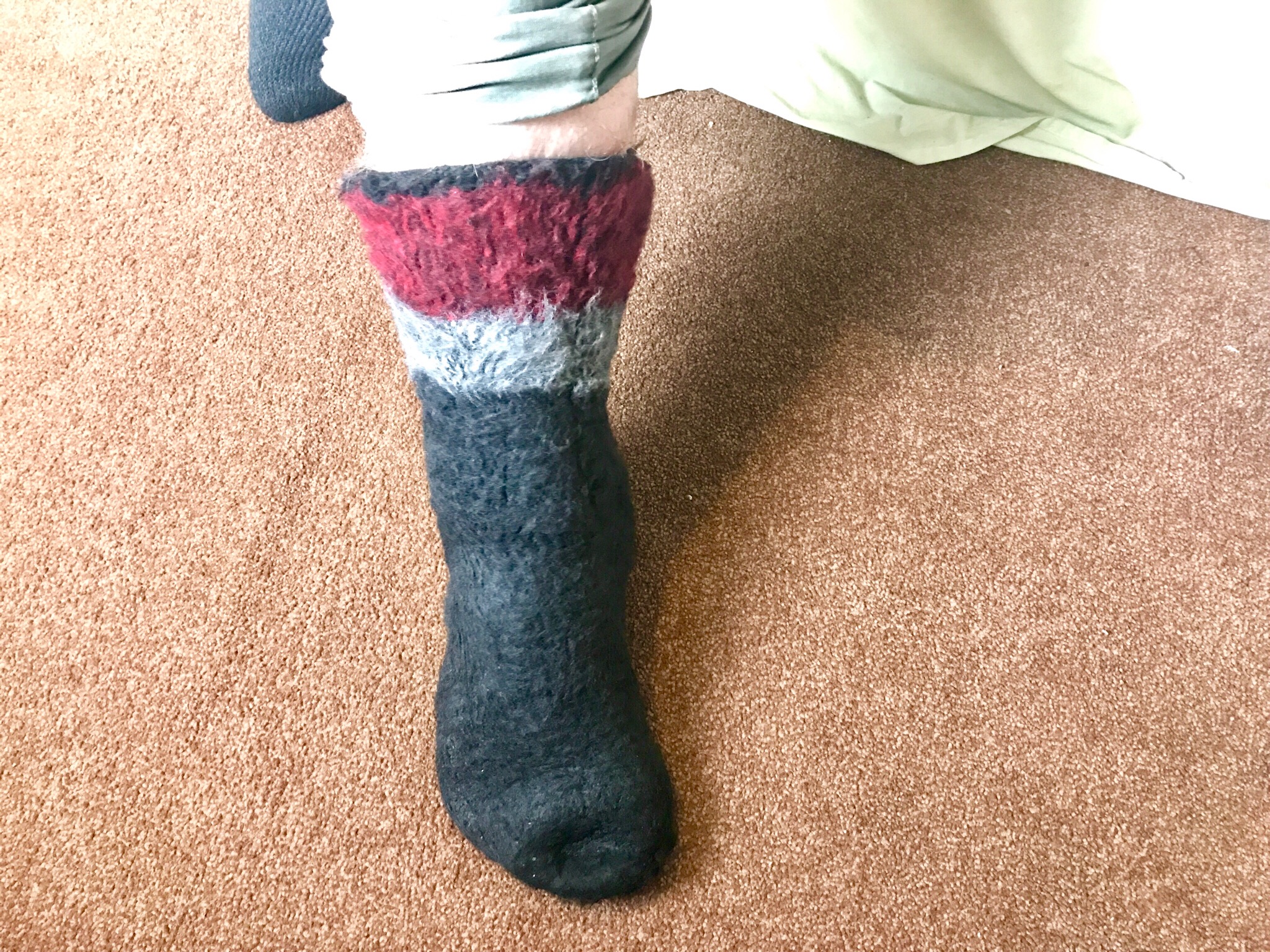 A person wearing a black sock that has a thick horizontal white stripe and a thinner bright Red stripe at the ankle, on a beige carpeted background.