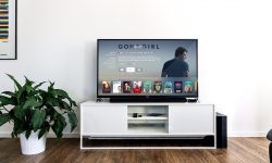 A clear glass TV Cabinet that has a wide black flat screen TV and a white vase full of some green flowers on it, on a white background.