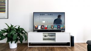 A clear glass TV Cabinet that has a wide black flat screen TV and a white vase full of some green flowers on it, on a white background.