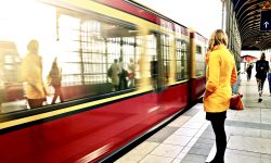 A large Red rectangular Train next to a stone platform with a medium height woman with blonde hair wearing a yellow coat, a black skirt, some black tights, and a pair of black heels and some various other people also stood on the platform, on a bright background.