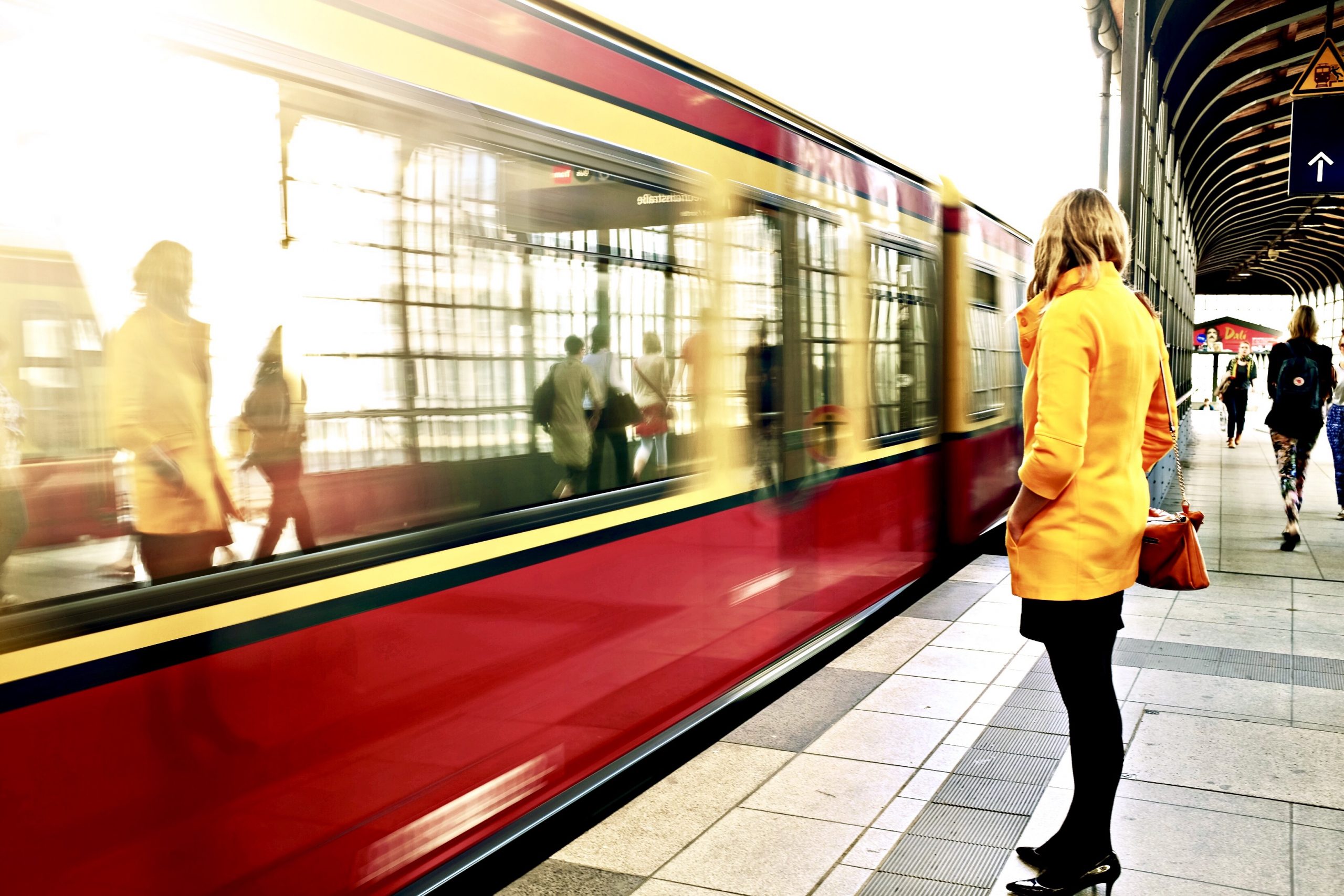 Five Things I hate About Travelling On Public Transport A large Red rectangular Train next to a stone platform with a medium height woman with blonde hair wearing a yellow coat, a black skirt, some black tights, and a pair of black heels and some various other people also stood on the platform, on a bright background.