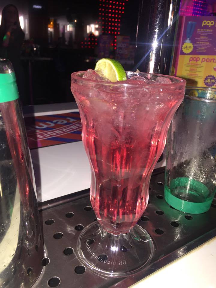 A large sundae glass full of some light pink coloured liquid, some clear ice cubes, some black straws, and a slice of Lemon on a metal tray, on a dark background.