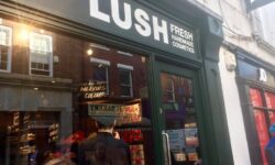 A black store front with a large glass window, some black boards, and a sign that has Lush written in large white writing and fresh handmade cosmetics written in smaller white writing on it, on a bright background.