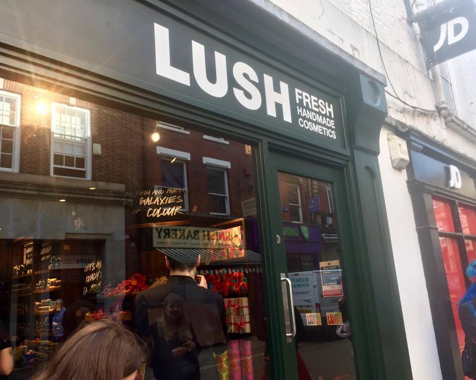 From Darkness To Light | Lush York Relaunch A black store front with a large glass window, some black boards, and a sign that has Lush written in large white writing and fresh handmade cosmetics written in smaller white writing on it, on a bright background.
