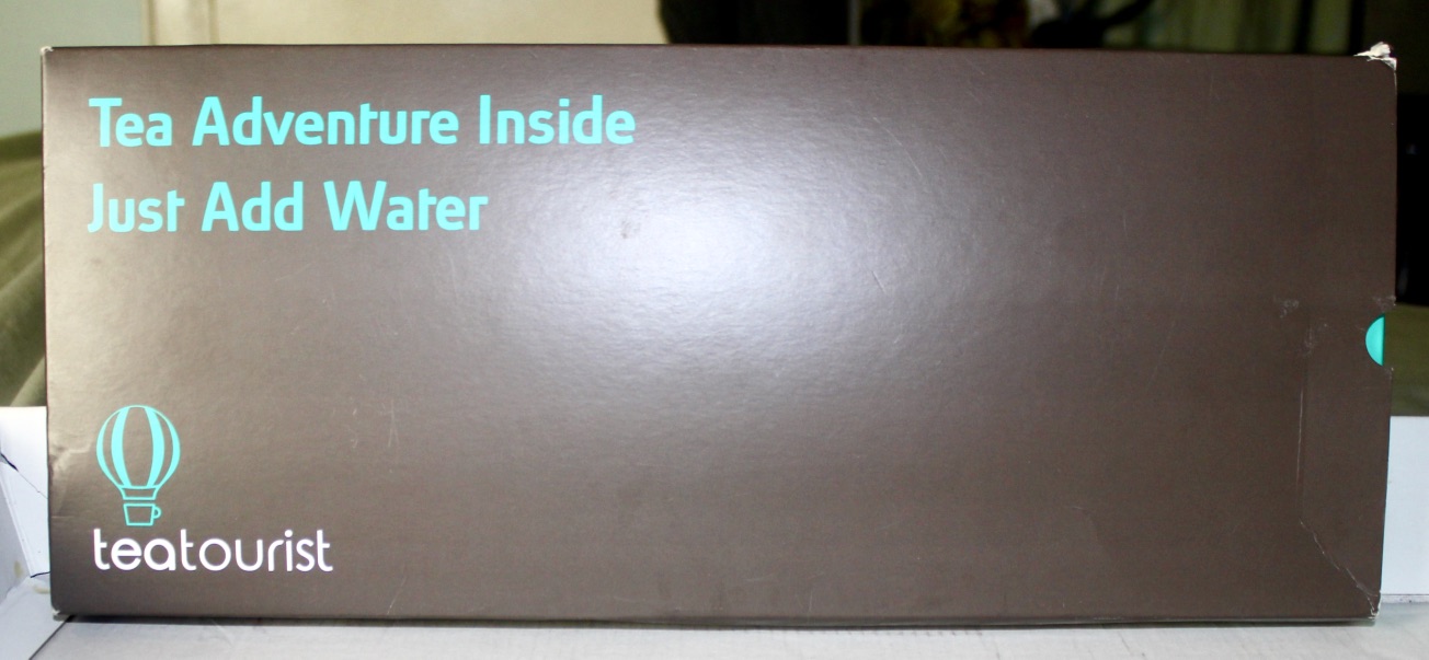 A rectangular black box that has Tea Adventure Inside Just Add Water written in Ice Blue writing and teatourist written in white writing on it, on a light background.