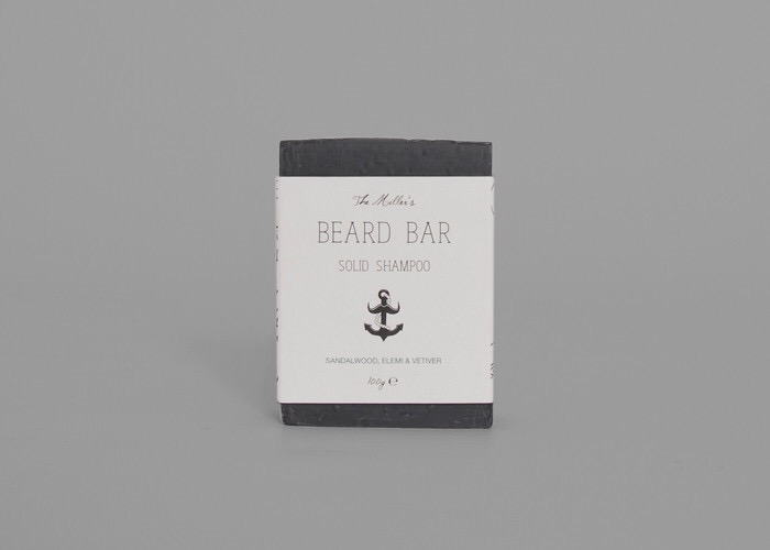 A rectangular black box covered with a white slip that has Brighton beard company beard bar written in black writing and a black anchor logo next to it, on a grey background.