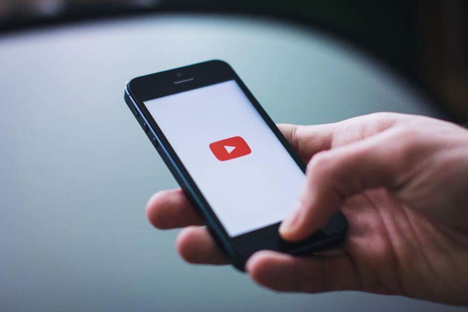 A white hand holding a black coloured smartphone that has a red coloured YouTube logo on it on a grey coloured marble table, on a dark background.