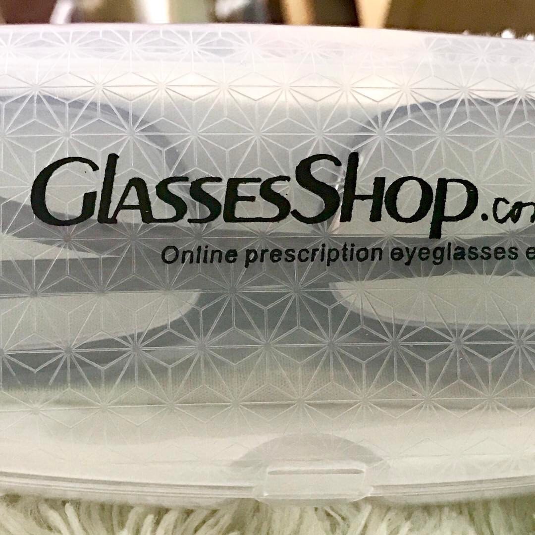 A pair of burgundy and silver coloured rectangular glasses inside a clear oval glasses case that has GlassesShop.com written in bold black writing on it, on a black background.