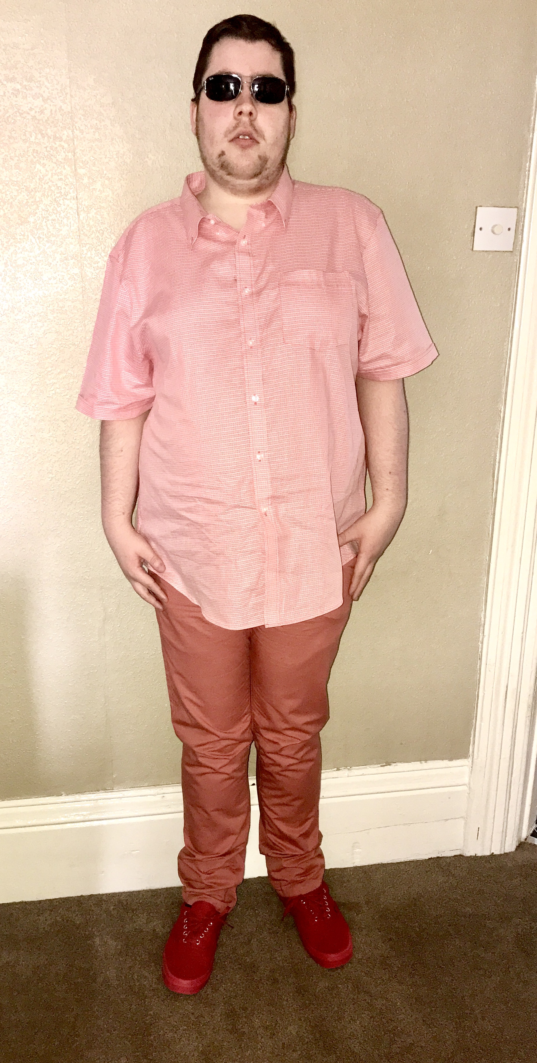 Me wearing a pair of dark green sunglasses, a short sleeved orange shirt, a pair of orange chinos, and a pair of red shoes with some red laces and soles next to a white wall, on a bright background.