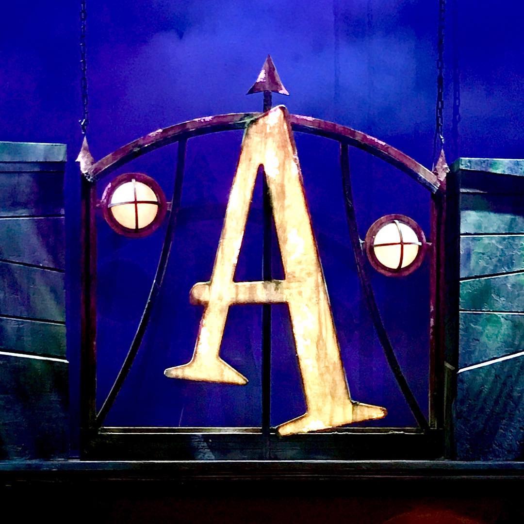 A large gold coloured Letter A, two circular lights, and an arch on a black platform, on a blue background.