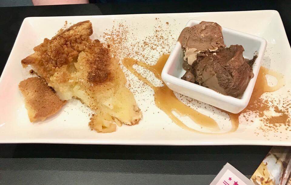 Some rectangular light brown pieces of pastry and light green pieces of Apple on a large rectangular white plate next to some chocolate ice cream with light brown biscuit pieces and caramel sauce in a small square white bowl on a black table, on a dark background.