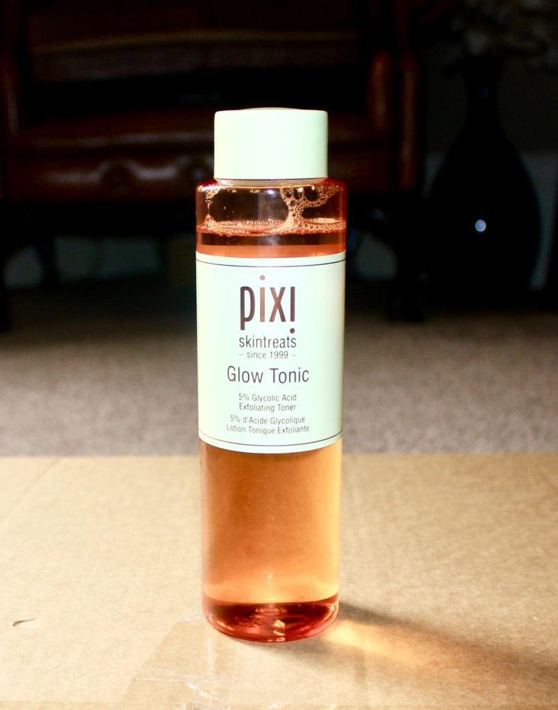The Tonic To Great Skin| Pixi Glow Tonic A tall slim cylindrical clear plastic bottle with a white lid full of some Orange liquid and a label that has Pixi Glow Tonic written in bold Grey writing on it on a light wooden table, on a black background.