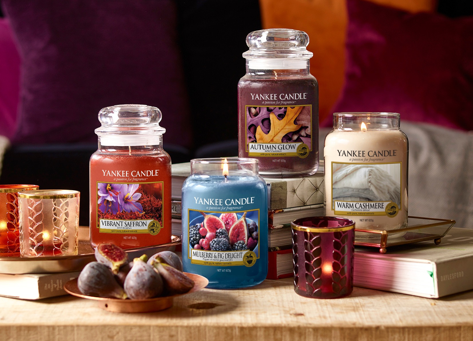 All four candles in some large glass jars next to some black and white votive holders containing different candles and a bowl of purple figs and mulberries on a light wood table, on a dark background.