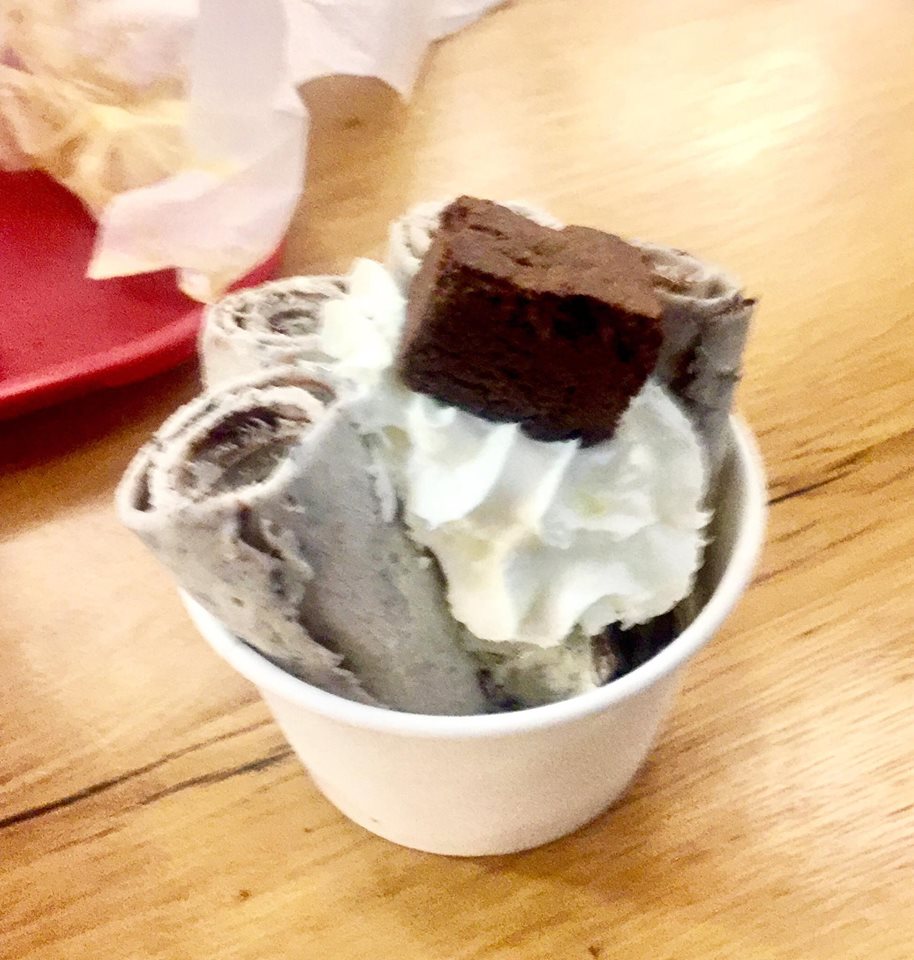 Some pale white cylindrical ice cream rolls with some black pieces of crushed Oreo, a dollop of white swirled whipped cream, and a deep brown rectangular chocolate brownie on top of it inside a circular white polystyrene bowl on a light wooden table, on a light background.