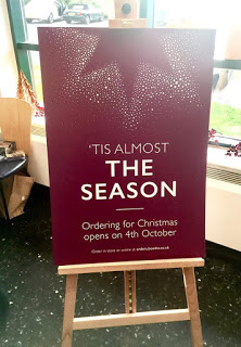 Filling Santa's Boots | Booths Ilkley Christmas 2017 Preview A dark Red Glittery advertisement board on a light wooden cardboard easel that has 'tis nearly written in small Gold writing and the season written in big bold white writing on it, on a nighttime background.