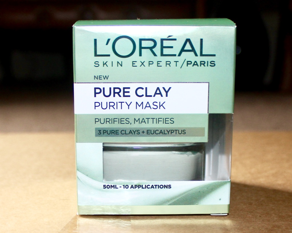 The Purity Of Great Skin | L'Oreal Paris Pure Clay Purity Face Mask A square silver coloured box that has L'Oreal written in large bold Grey writing, Paris written in smaller Grey writing, and Pure Clay written in bold dark grey writing on a wooden table, on a bright background.