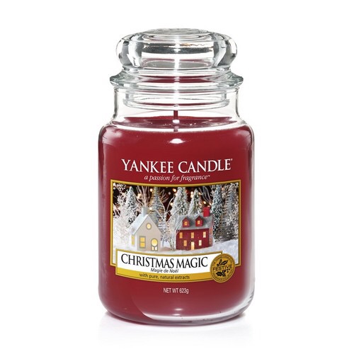 A tall glass jar full of some bright Red coloured wax with a label that has yankee candle written in white writing, Christmas Magic written in black writing, and a picture of some trees and some pale golden houses on it, on a white background.