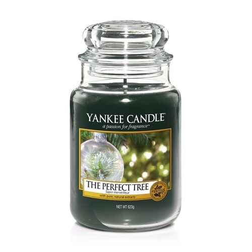 A tall glass jar full of some dark brown coloured wax with a label that has yankee candle written in white writing, The Perfect Tree written in black writing, and a picture of a tree inside a snow globe with some twinkling fairy lights on it, on a white background.