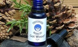 A tall black cylindrical bottle with a pipette lid and white label that has ThatchFace written in large bold black writing, Got Wood Beard Oil written in smaller black writing, and a picture of a blank face with a white beard and moustache inside a green triangle on it, on a light background.