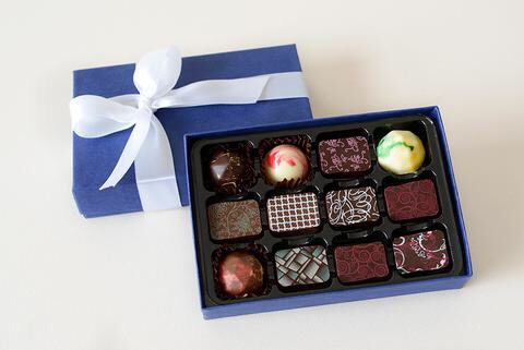 Luke Loves Christmas 2017 | Mens Under £50 Gift Guide A large rectangular landscape box full of some circular and rectangular black and white chocolates next to a Purple box lid and a large white tied ribbon, on a white background.