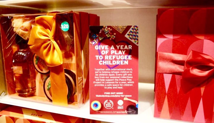 Peace On Earth | The Body Shop Leeds Briggate Christmas 2017 Preview Two tall red gift boxes with a red and gold tied ribbon on the top of them sandwiched between a bright red card that has Give A Year Of Play To Refugee Children written in bright bold white writing on it on a rectangular clear glass shelf, on a bright background.