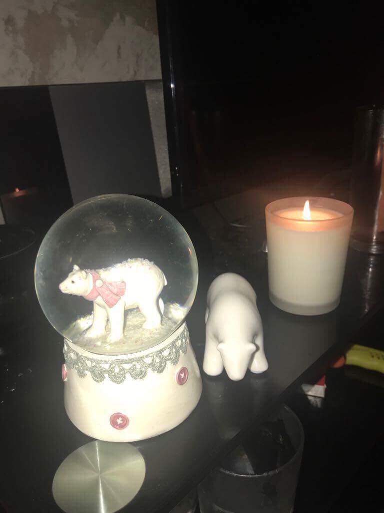 Luke Loves Christmas 2017 | How I've Decorated My Room This Year A large bright white Snow Globe that has a Polar Bear stood on all fours inside it next to a small white Polar Bear ornament also stood on all fours and a tall white burning candle in a clear glass jar on a glass cabinet, on a dark background.