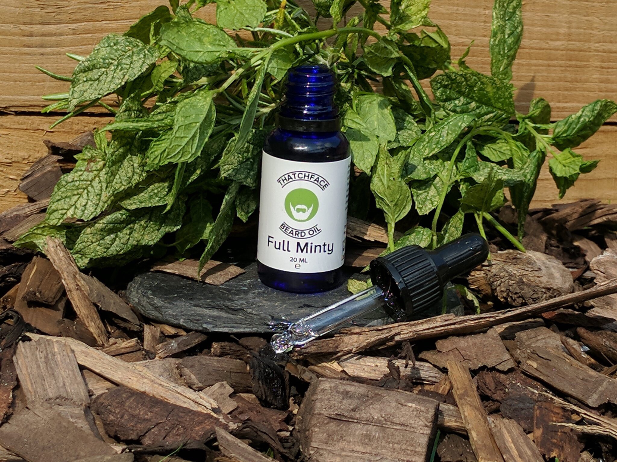 A tall black cylindrical dropper bottle with a white label that has Thatchface written in large bold black writing, Full Minty Beard Oil written in smaller black writing, and a picture of a blank face with a white beard and moustache inside a green triangle, on a light background.