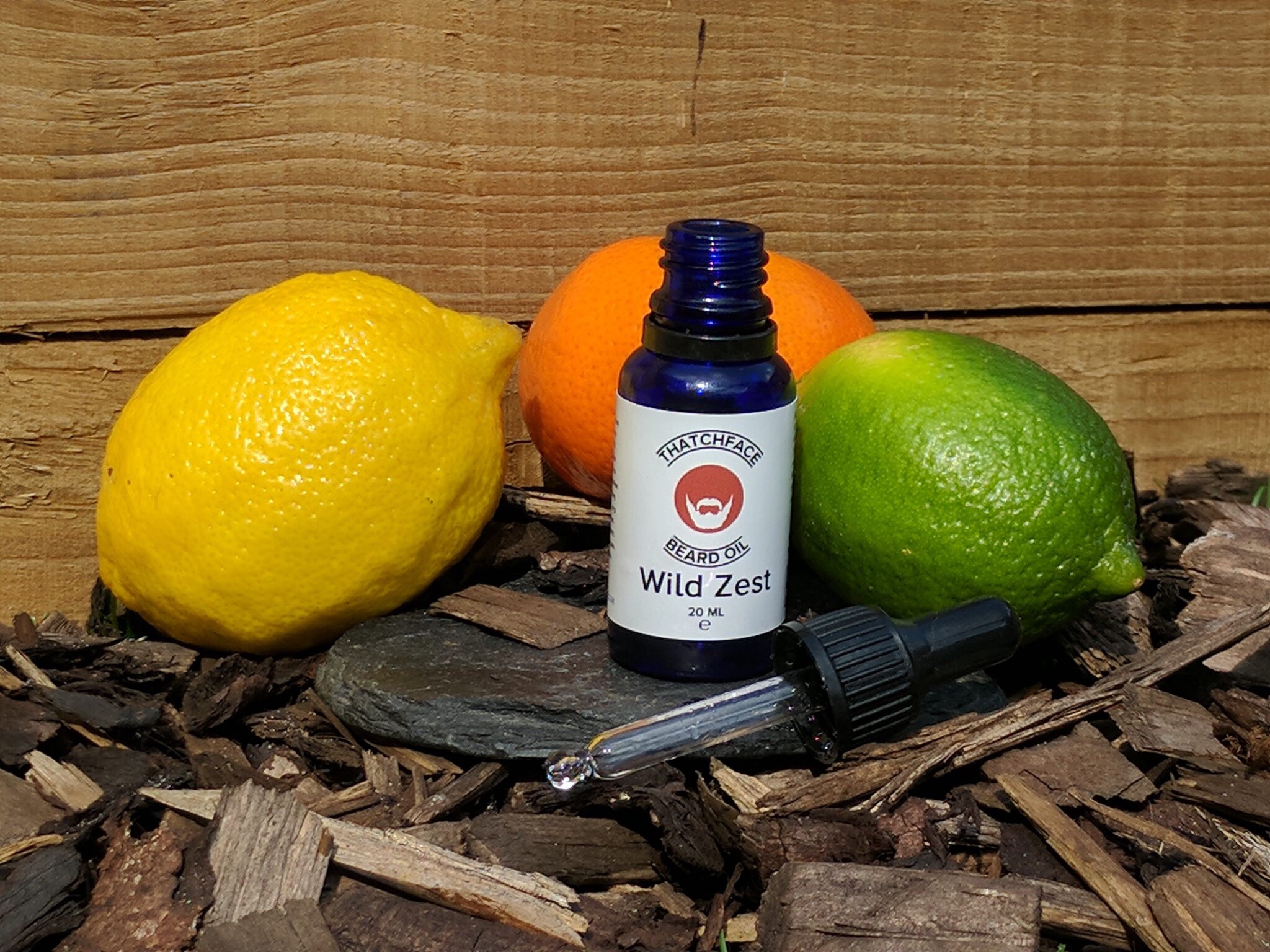 A tall black cylindrical dropper bottle with a white label that has Thatchface written in large bold black writing, Wild Zest Beard Oil written in smaller black writing, and a picture of a blank face with a white beard and moustache inside a green triangle, on a light background.