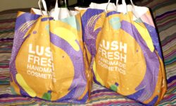 Two large paper bags that have some different bright swirling colours on the front of them on top of a stripy duvet cover, on a white background.