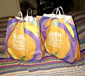 Two large paper bags that have some different bright swirling colours on the front of them on top of a stripy duvet cover, on a white background.