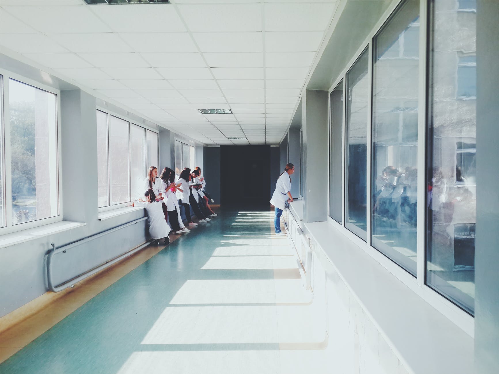 A long white corridor full of some light wooden doors, some large light wooden tables, and some bright plastic chairs, on a light background.