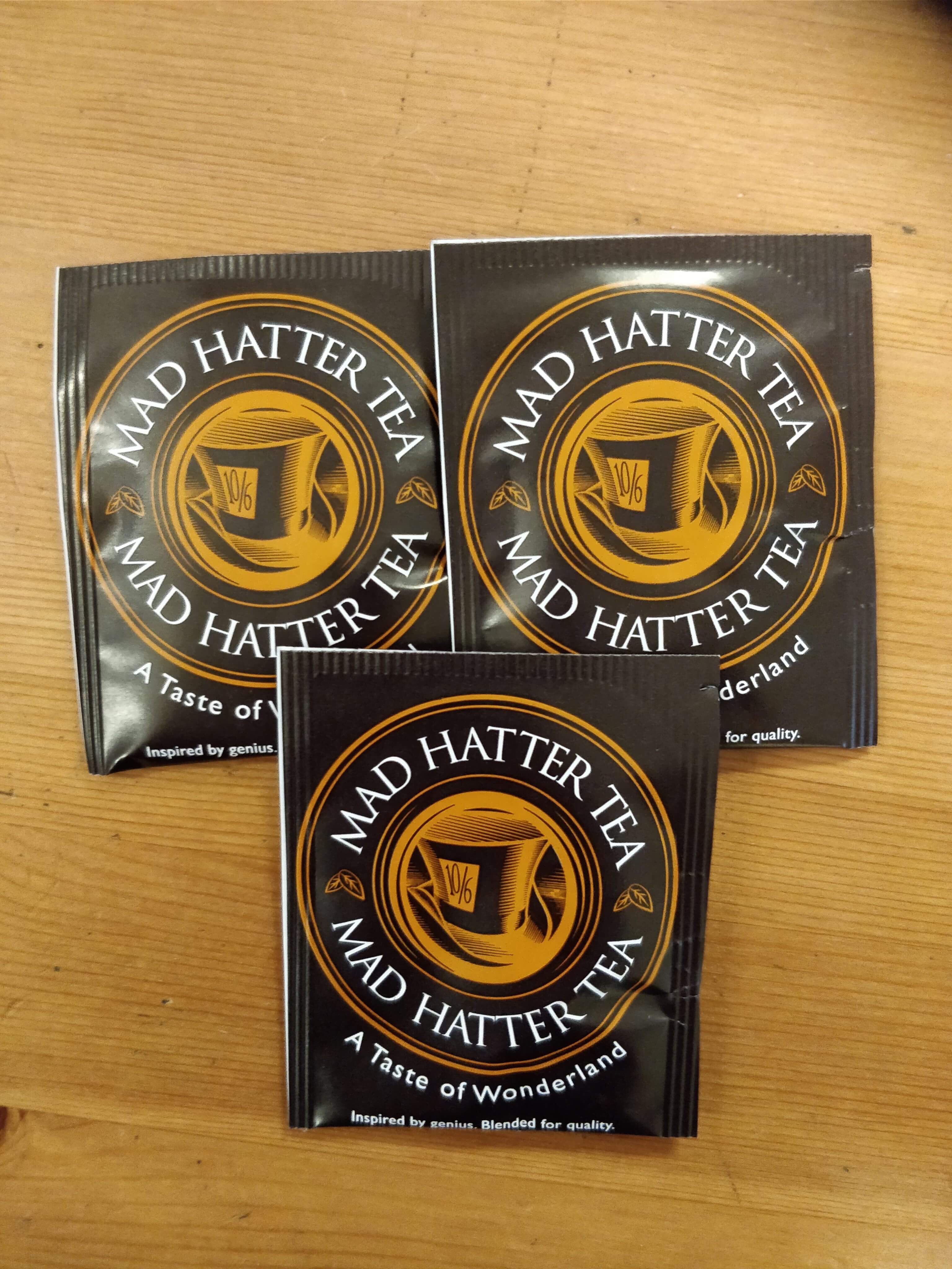Tea Tastes Terrific | The Mad hatter Tea Company Original Tea Bags Three black square paper pouches that have a circular logo with a gold tea cup, the mad hatter tea company written in bold white writing, and a taste of wonderland written in smaller white writing on them, on a light brown background.