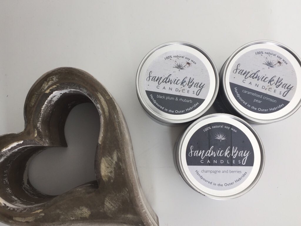 Great Scent Always Travels Well | Sandwick Bay Candles Small Travel Tin Candles three small silver metal jars full of some white coloured wax with some white labels that have Sandwick Bay Candles Caramelised Crimson Pear, Black Plum & Rhubarb, and Champagne & Berries written in bold grey writing on them next to a ceramic brown heart ornament, on a light grey background.
