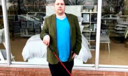 me stood on some grey stone paving in front of a large rectangular window wearing a medium green jacket, a bright blue jumper, a pair of black trousers, and a pair of black Leather boots holding a long red cane, on a light background.