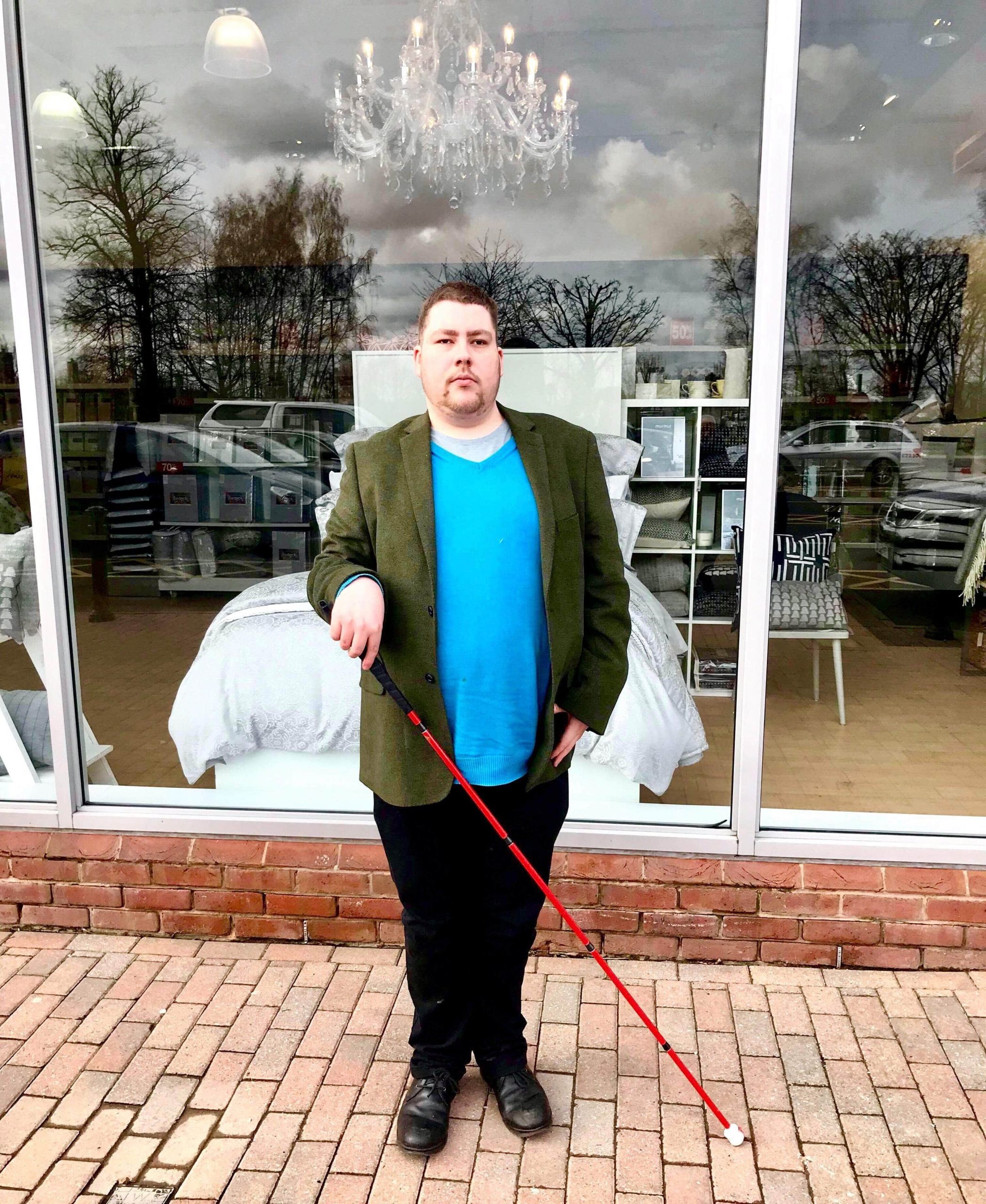 me stood on some grey stone paving in front of a large rectangular window wearing a medium green jacket, a bright blue jumper, a pair of black trousers, and a pair of black Leather boots holding a long red cane, on a light background.