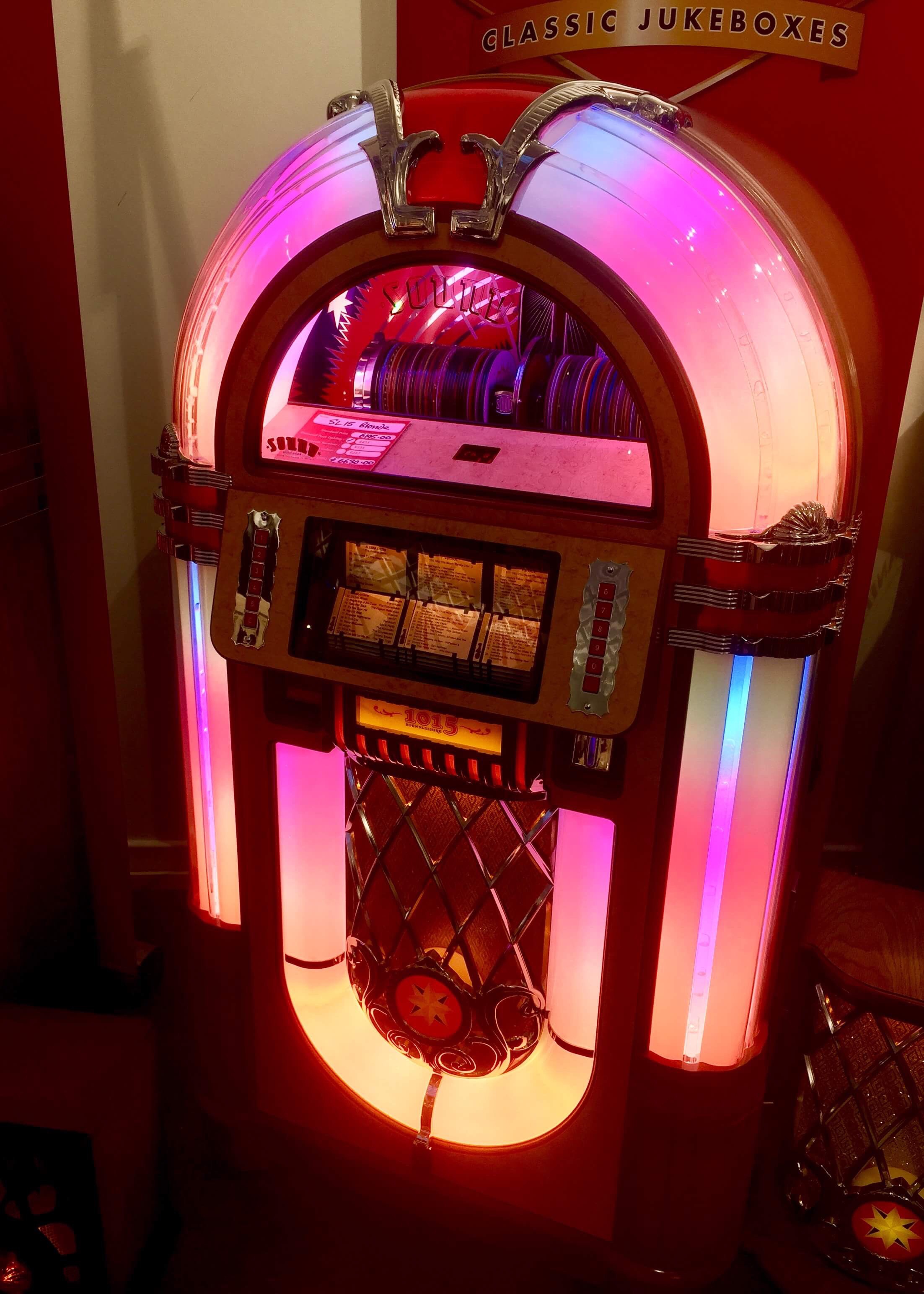 An arch shaped clear glass and dark brown wooden Jukebox that has a semi circled clear glass screen, some white plastic square buttons, and some yellow, pink and purple lights on it, on a dark background.