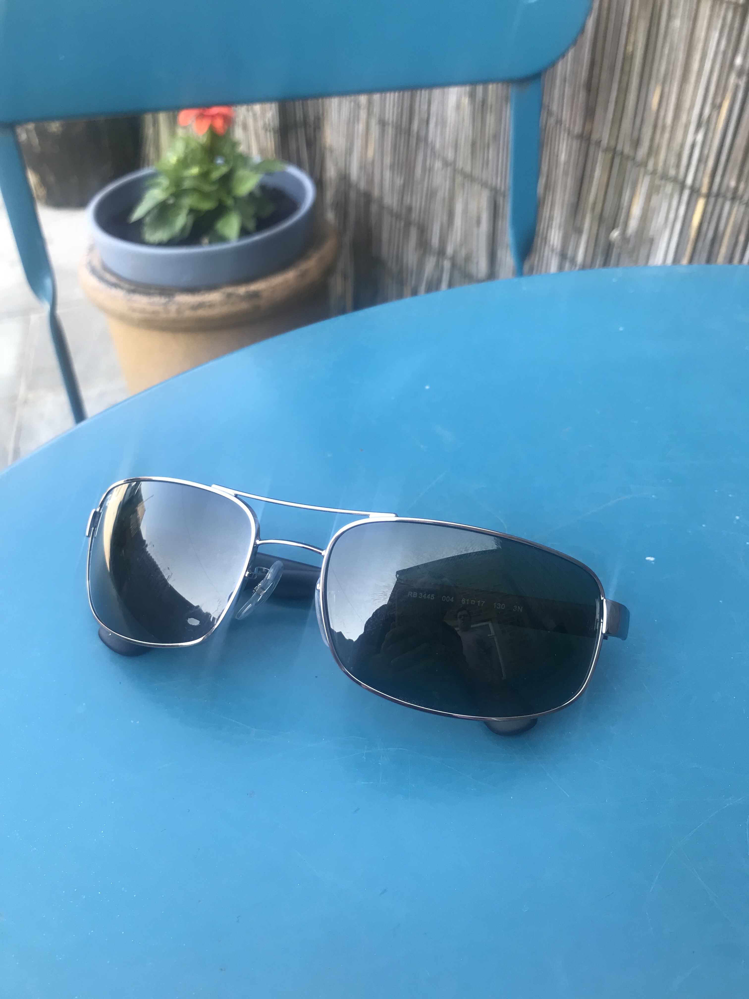A pair of folded Sunglasses with black plastic arms, a Silver Metal frame and dark Green Lenses on top of a blue garden table, on a white background.