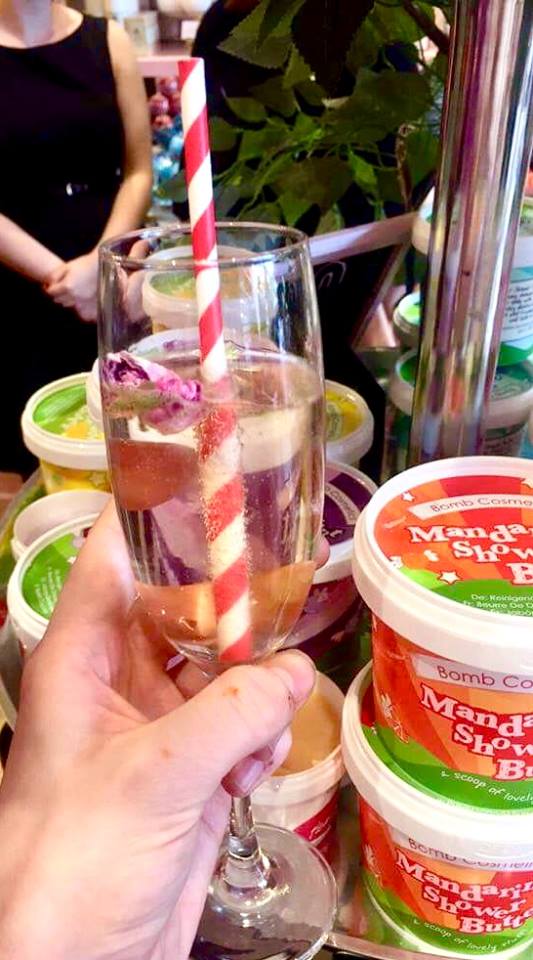 A tall clear glass champagne flute full of some clear liquid and some pale red rose petals floating inside it with a cylindrical white and red paper straw, on a bright background.