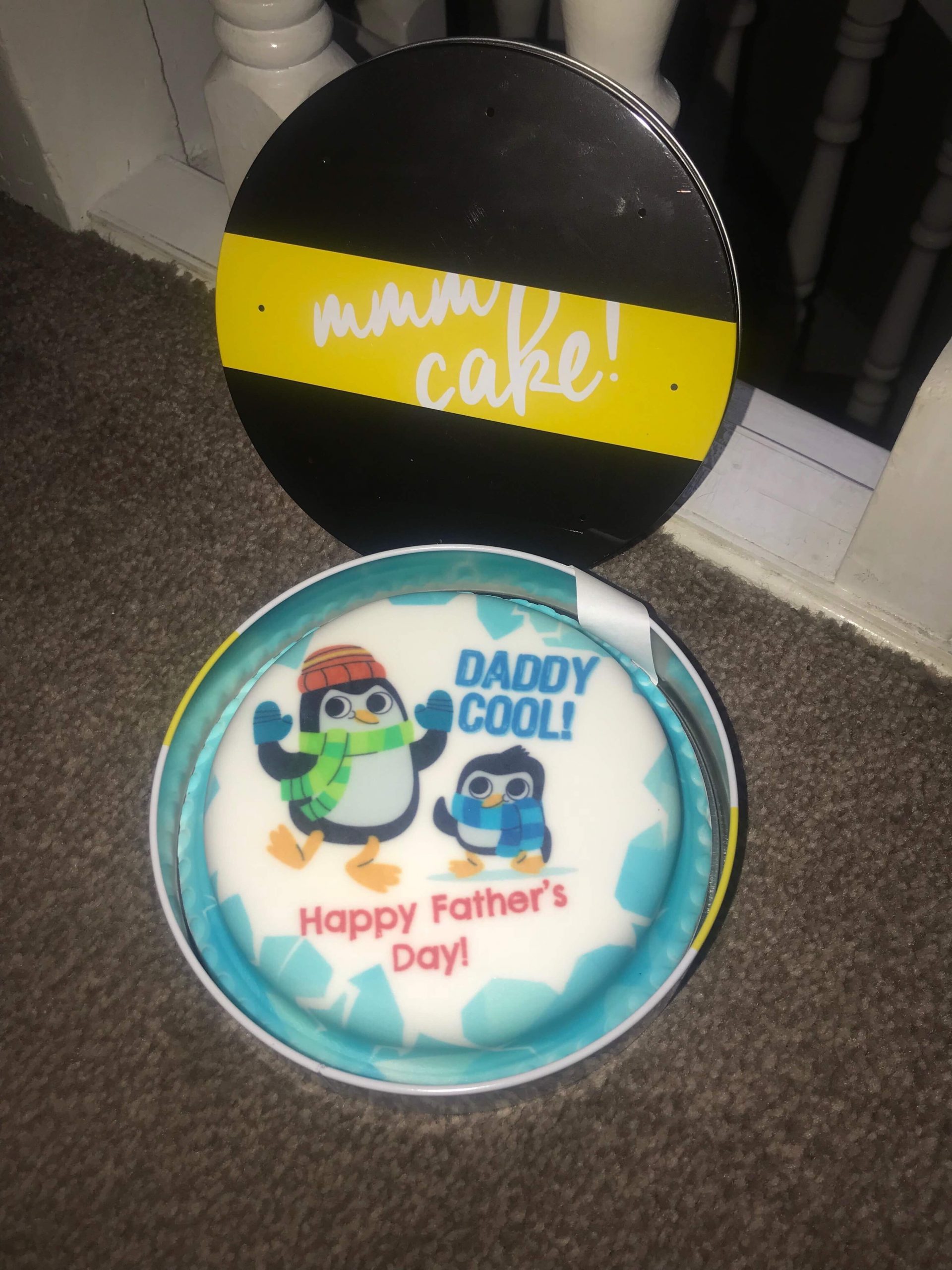 Cake! Cake! I Just Love Cake! | Bakerdays Personalised Father's Day Lemon Drizzle Letterbox Cake a black and yellow metal cake tin full of some white and blue coloured cake that has daddy cool written in blue icing, happy fathers day written in red icing and a picture of two black and white penguins wearing some hats and scarfs on it, on a white background.