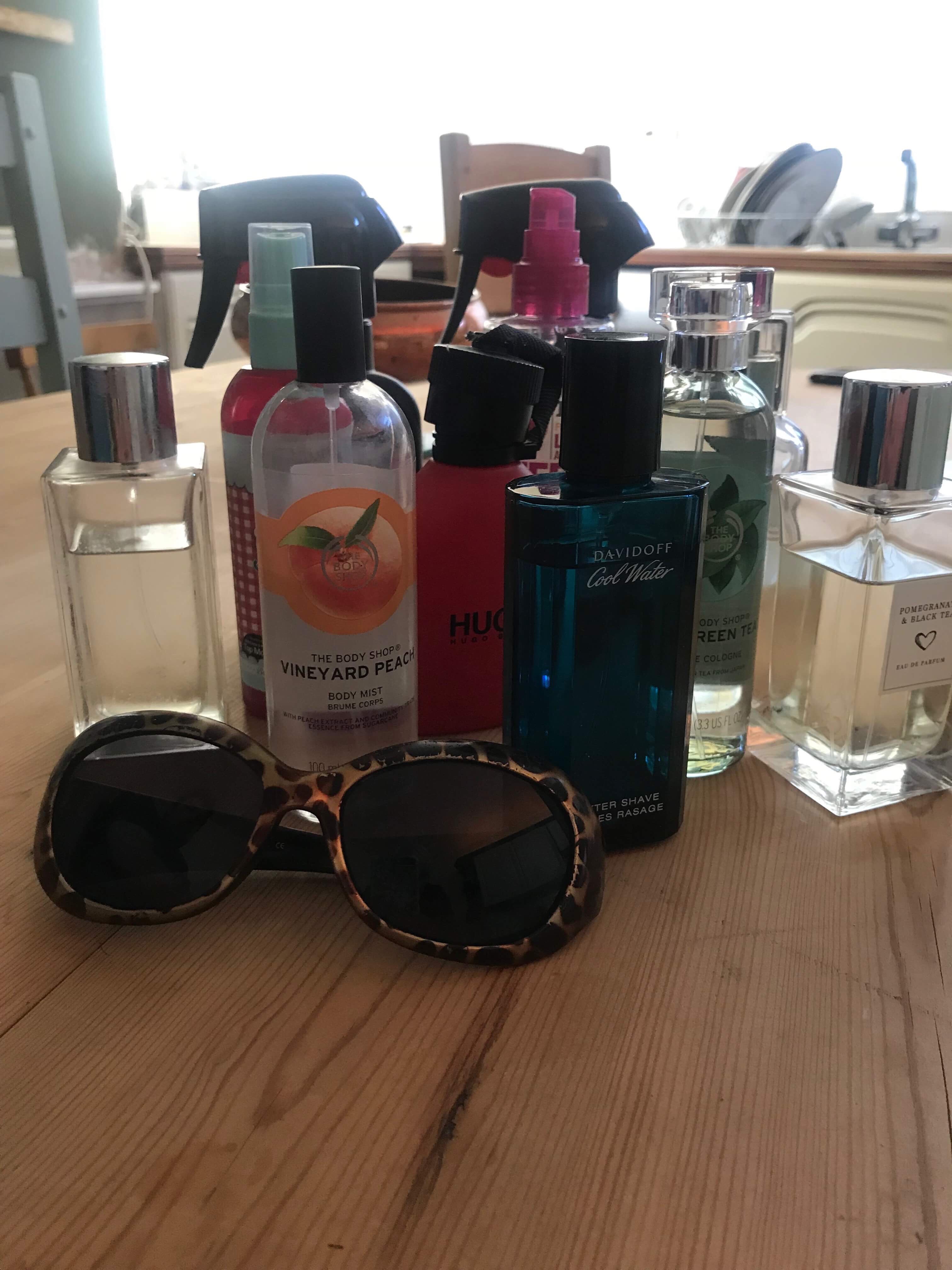 Smelling As Fresh As A Daisy | What's On My Fragrance Shelf July 2018 A variety of tall rectangular glass bottles, cylindrical glass bottles, tall plastic kitchen spray bottles, and tall cylindrical plastic bottles on a wooden table, on a white background.