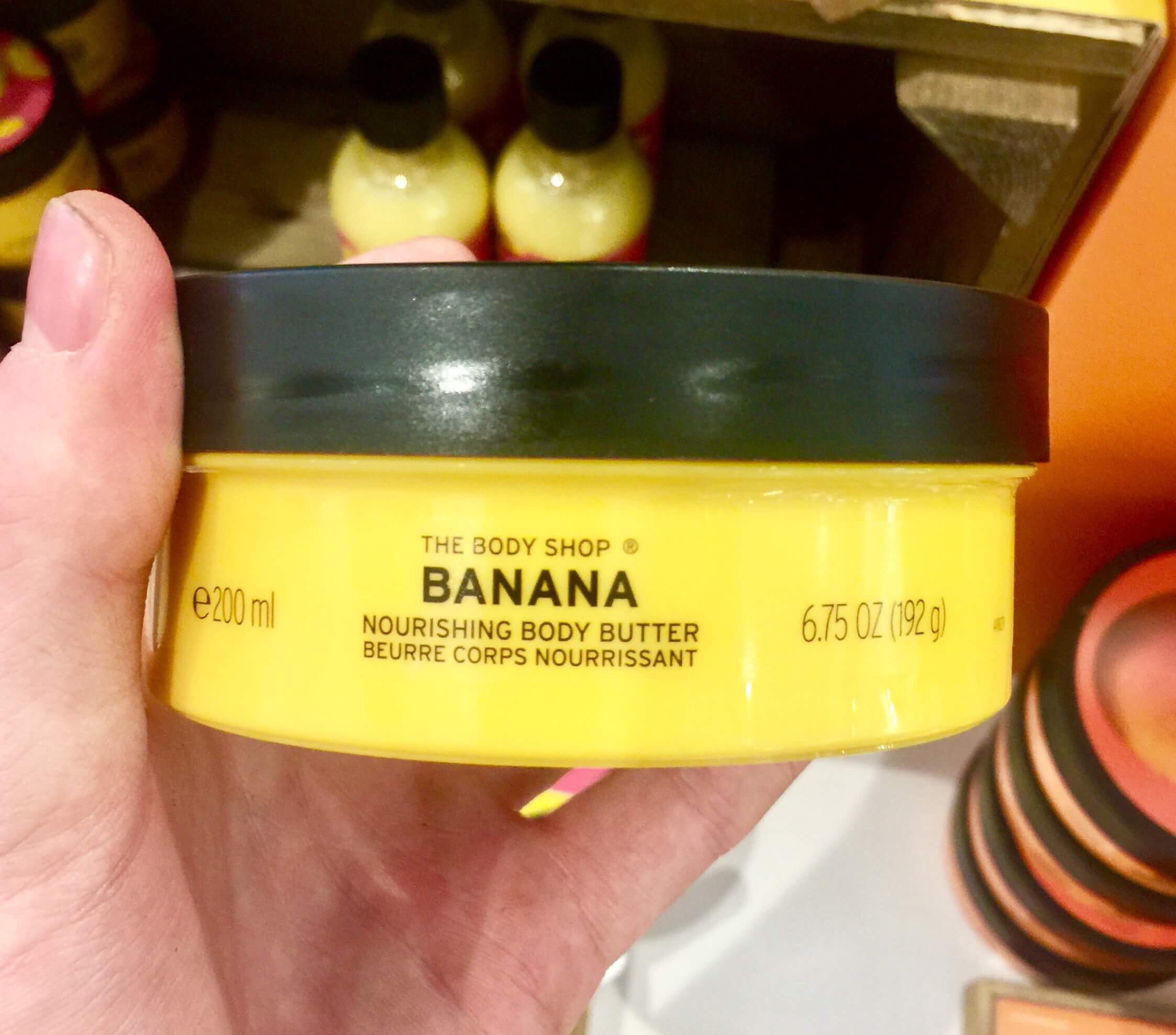 A short cylindrical yellow plastic tub with a black lid that has The Body Shop Banana Body Butter written in bold black writing and some pictures of some Bananas on it, on a white background.