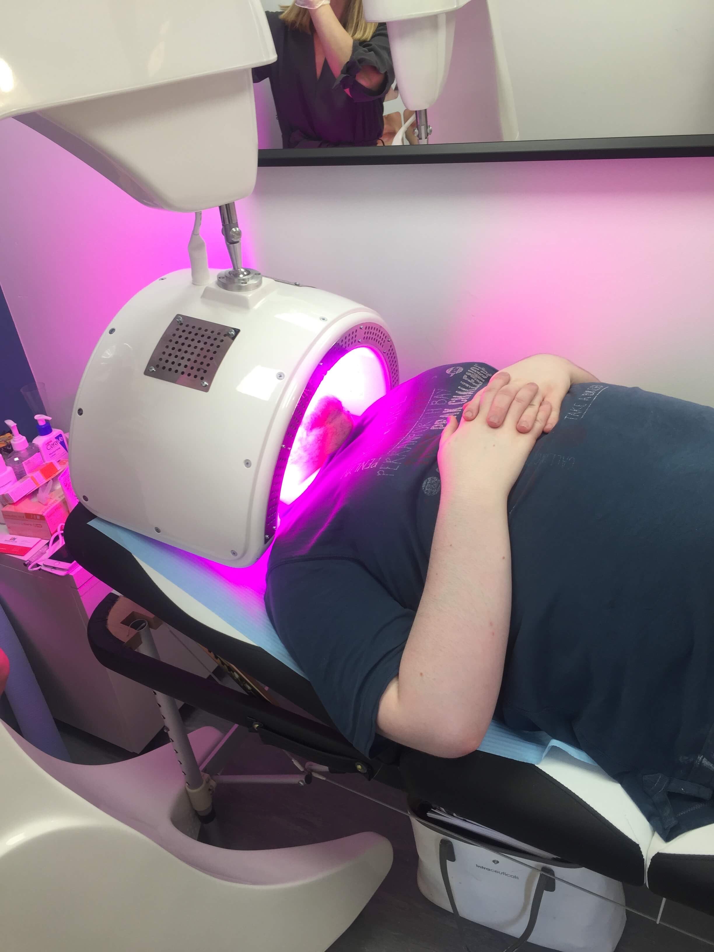 The Tingle Of Youth | GlamGlow YouthMud® Tinglexfoliate Treatment Me laid on a white medical bed wearing a black t-shirt with a white plastic semi-circle light panel over my Face, on a white background.