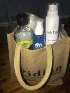 A light Green rectangular hessian tote Bag full of some different shaped plastic bottles and some glass jars, on a white background.