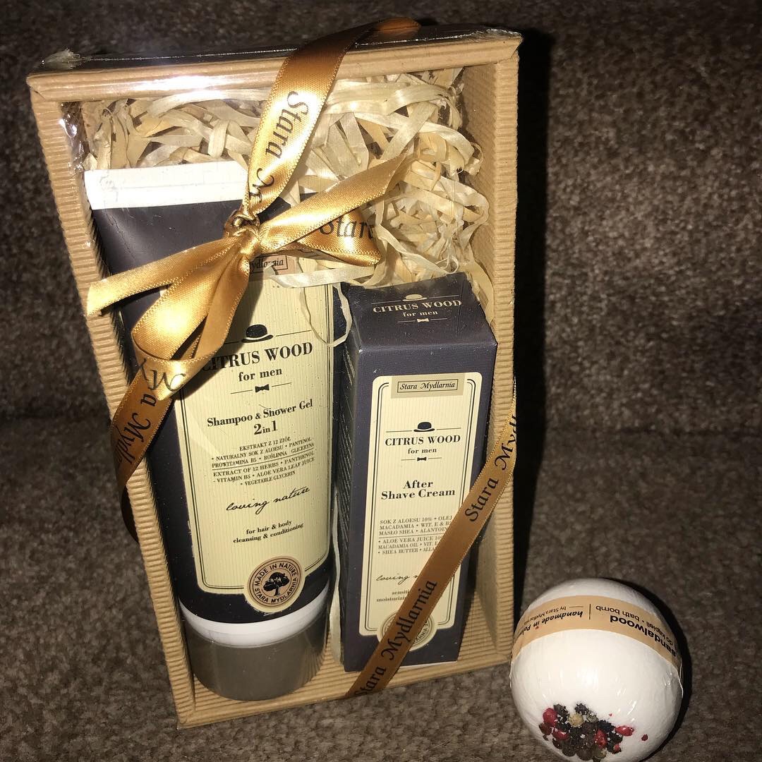 A spherical white bath bomb with a white label that has Sandalwood written in bold black writing in front of a tall cone shaped Silver plastic bottle that has True Obsession Citrus Wood 2 in 1 Shower Gel + Shampoo written in bold black writing and a short rectangular Cardboard box that has True Obsession Mens Citrus Wood Aftershave Cream written in bold black writing on it inside a large cardboard box full of some hay, on a white background.