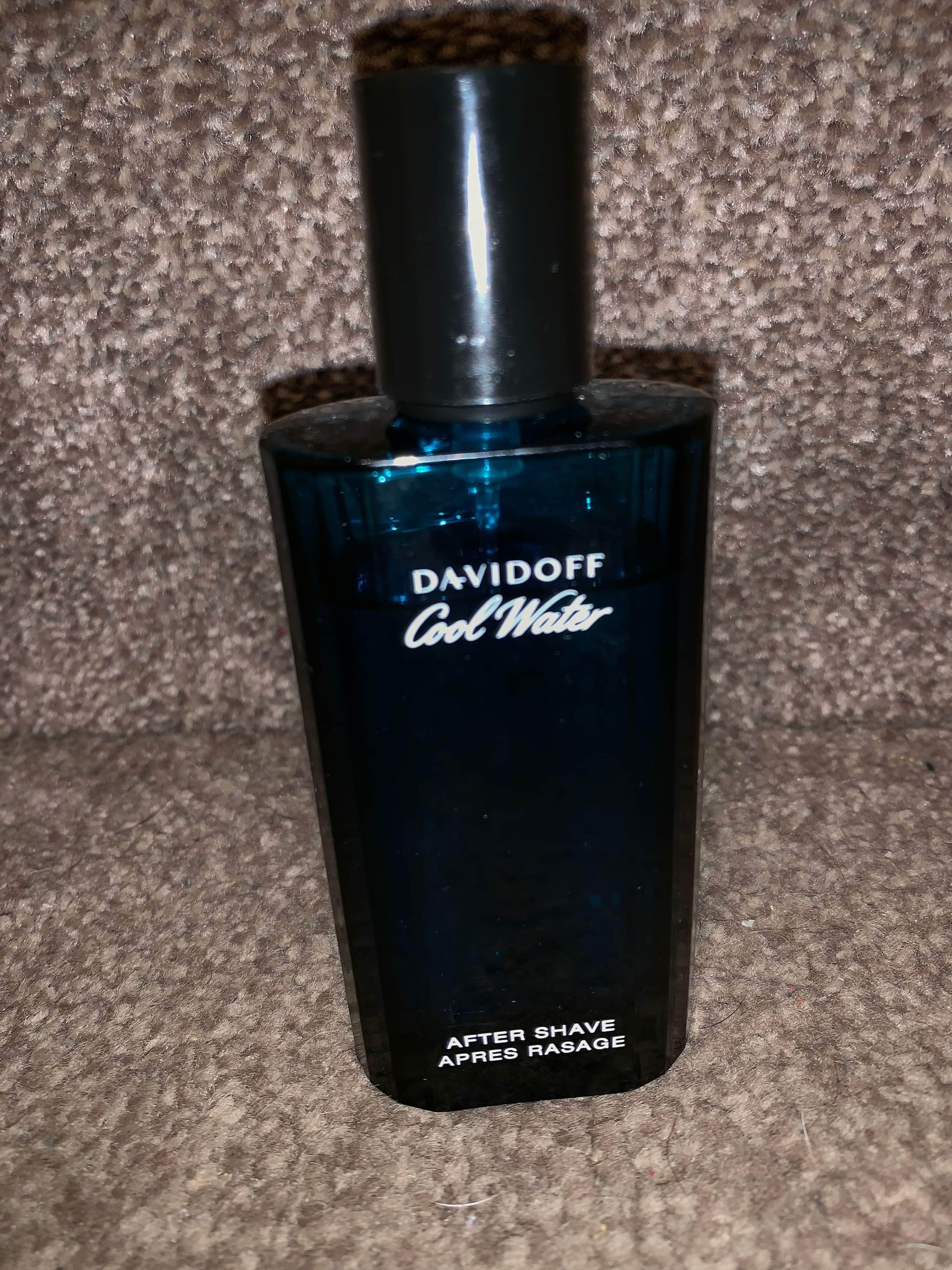 A tall rectangular blue glass bottle full of some clear liquid with a silver metal lid that has Davidoff Cool Water Man Eau De Toilette written in bold white writing on it, on a white background.
